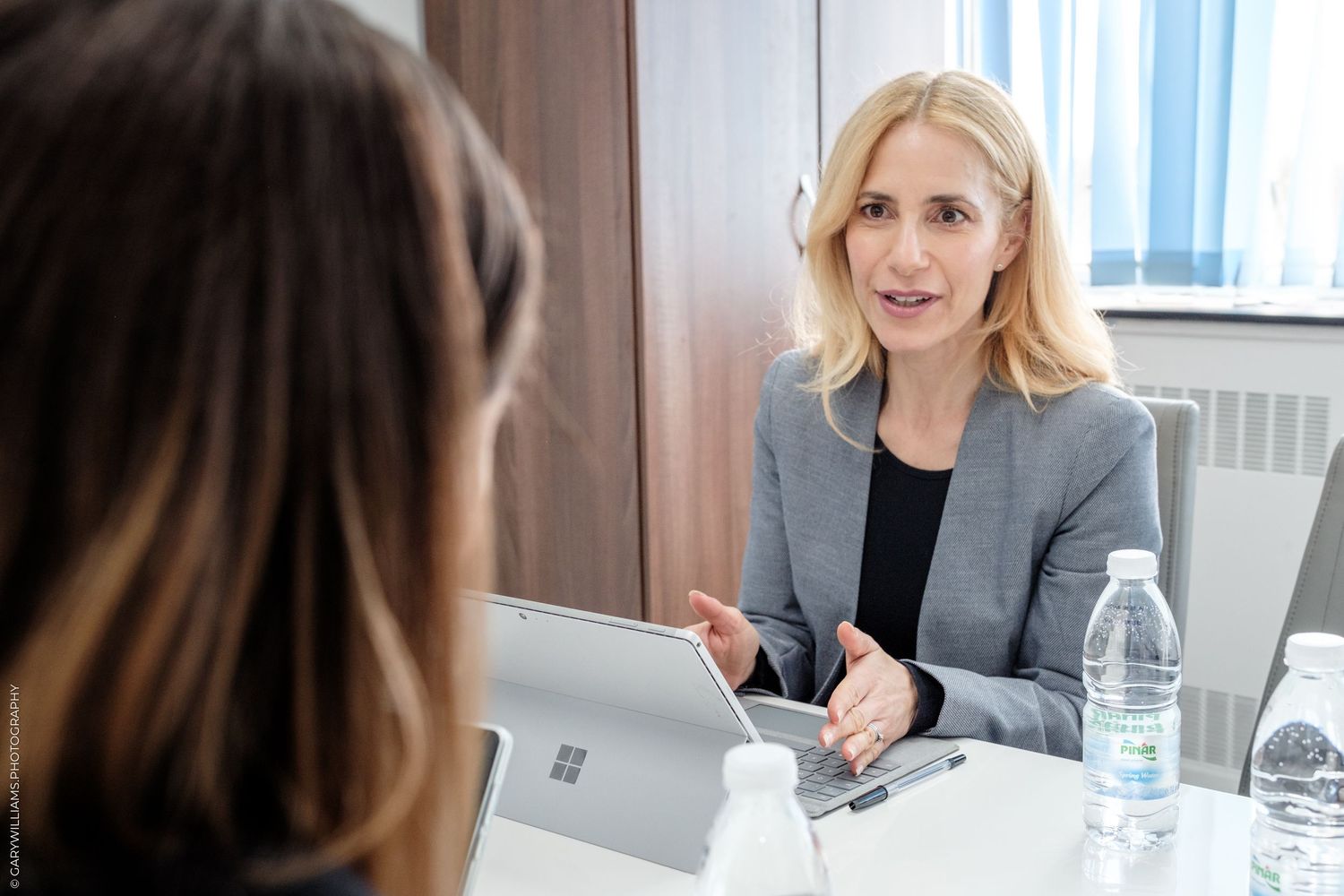 Professional businesswoman in gray blazer discussing strategy with colleague over Microsoft Surface tablet in modern office.