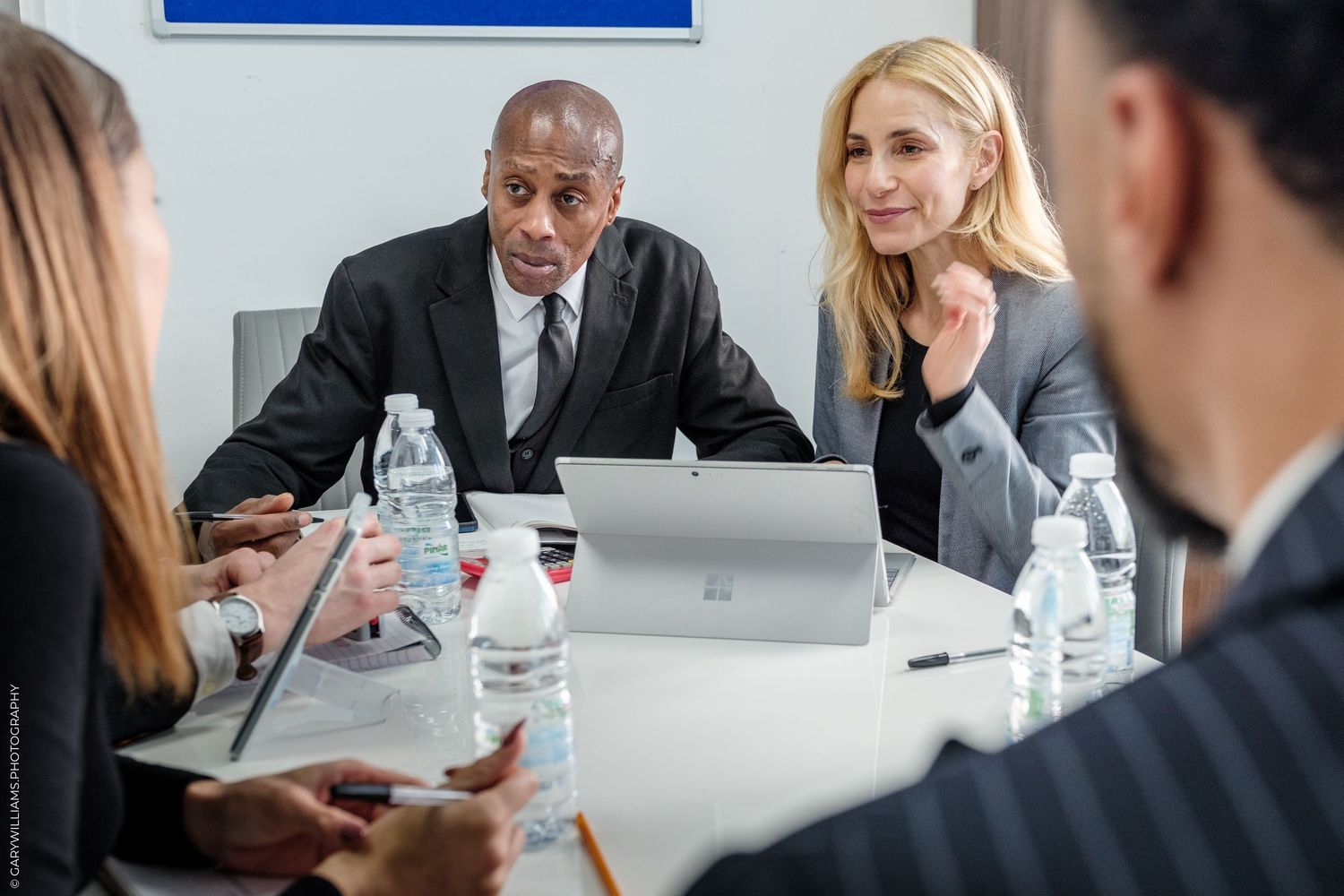 Business professionals in suits having a meeting around a conference table with laptops and water bottles.