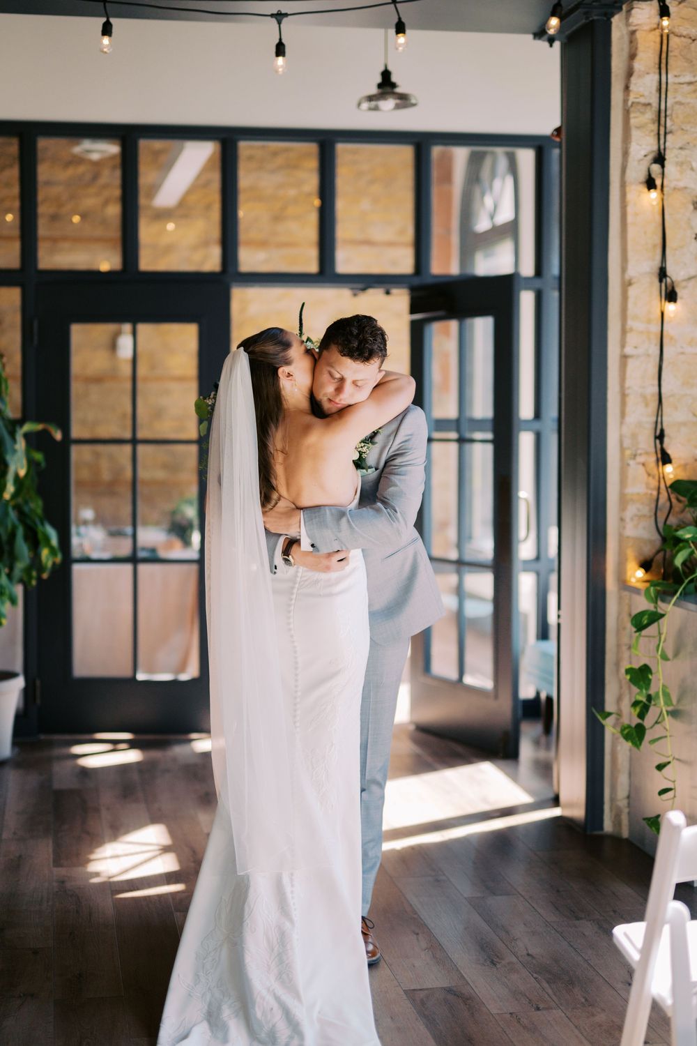 A couple embraces in a modern indoor venue with string lights and large windows casting natural light.
