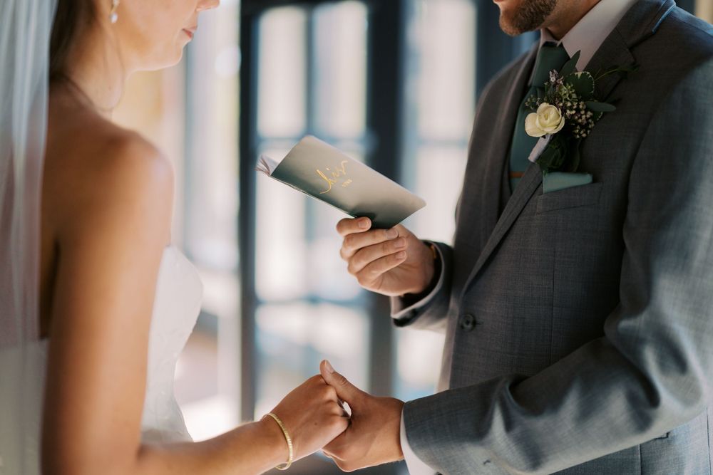 A couple holds hands during a wedding ceremony as vows are read from a small book in natural window light.