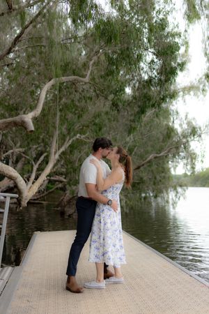 A romantic scene unfolds on a wooden dock surrounded by willow trees hanging over calm water.