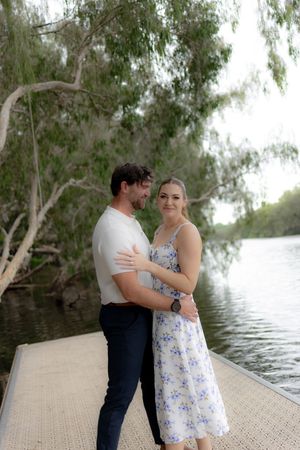 Two people share an intimate moment on a dock by a serene lake surrounded by willow trees.