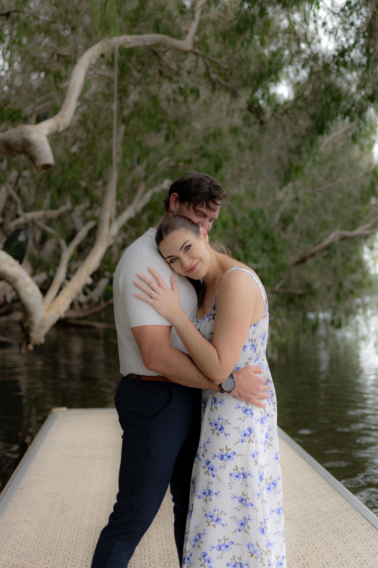 A couple shares an intimate moment on a wooden dock surrounded by trees and water.