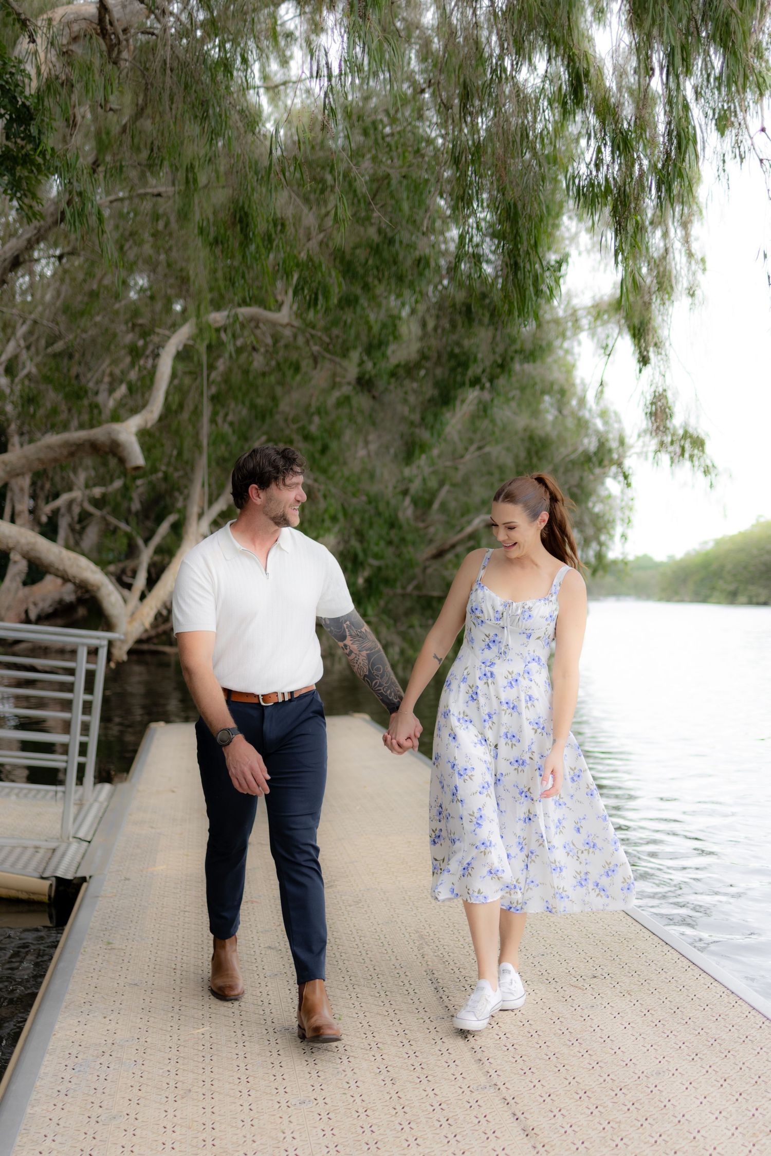 Couple walking hand in hand along a wooden dock by a lake with willow trees creating a romantic atmosphere.