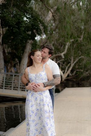 A couple embraces on a wooden dock by the water, with the woman wearing a floral white and blue sundress.