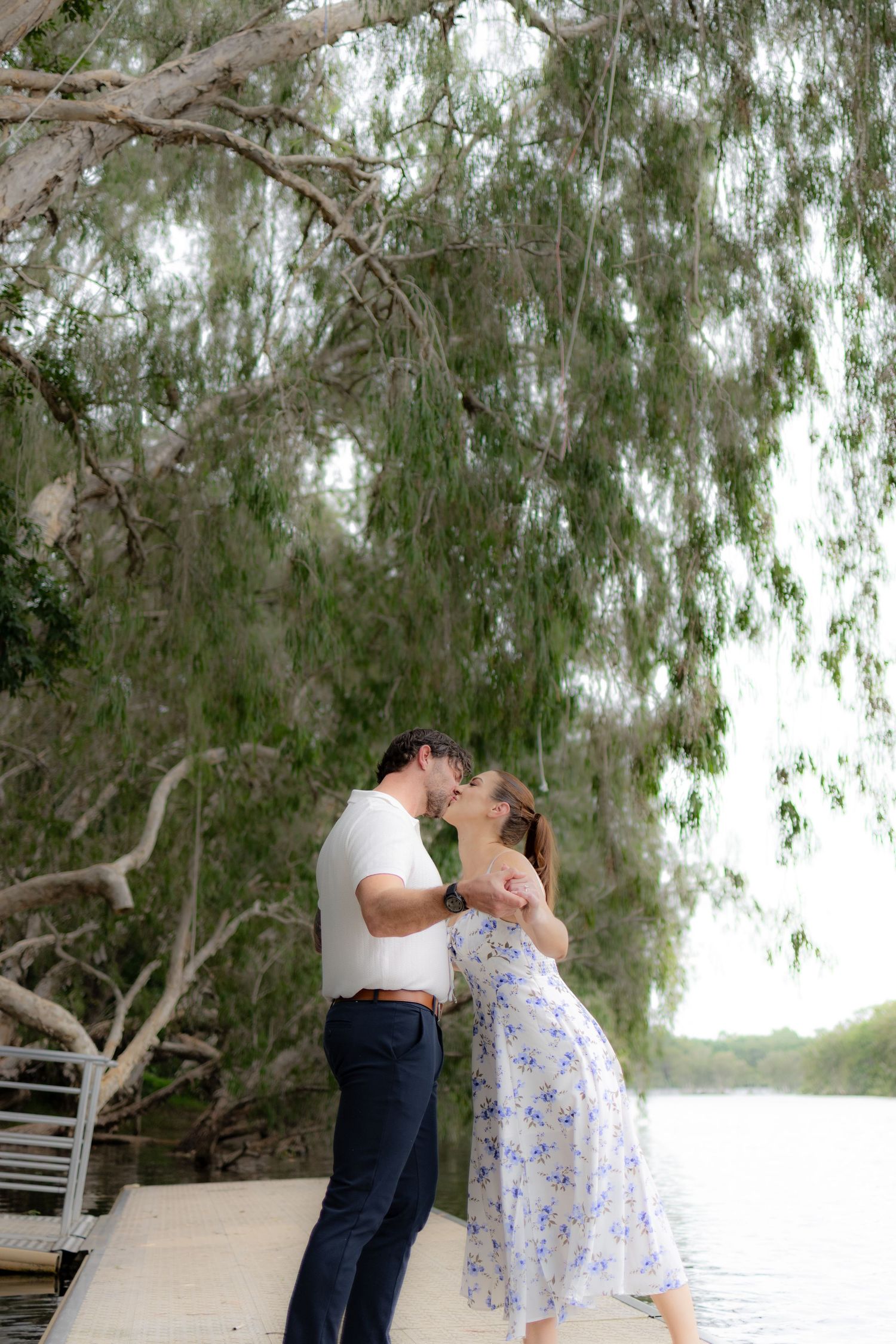A romantic couple shares tender moments under willow trees in a scenic outdoor setting.