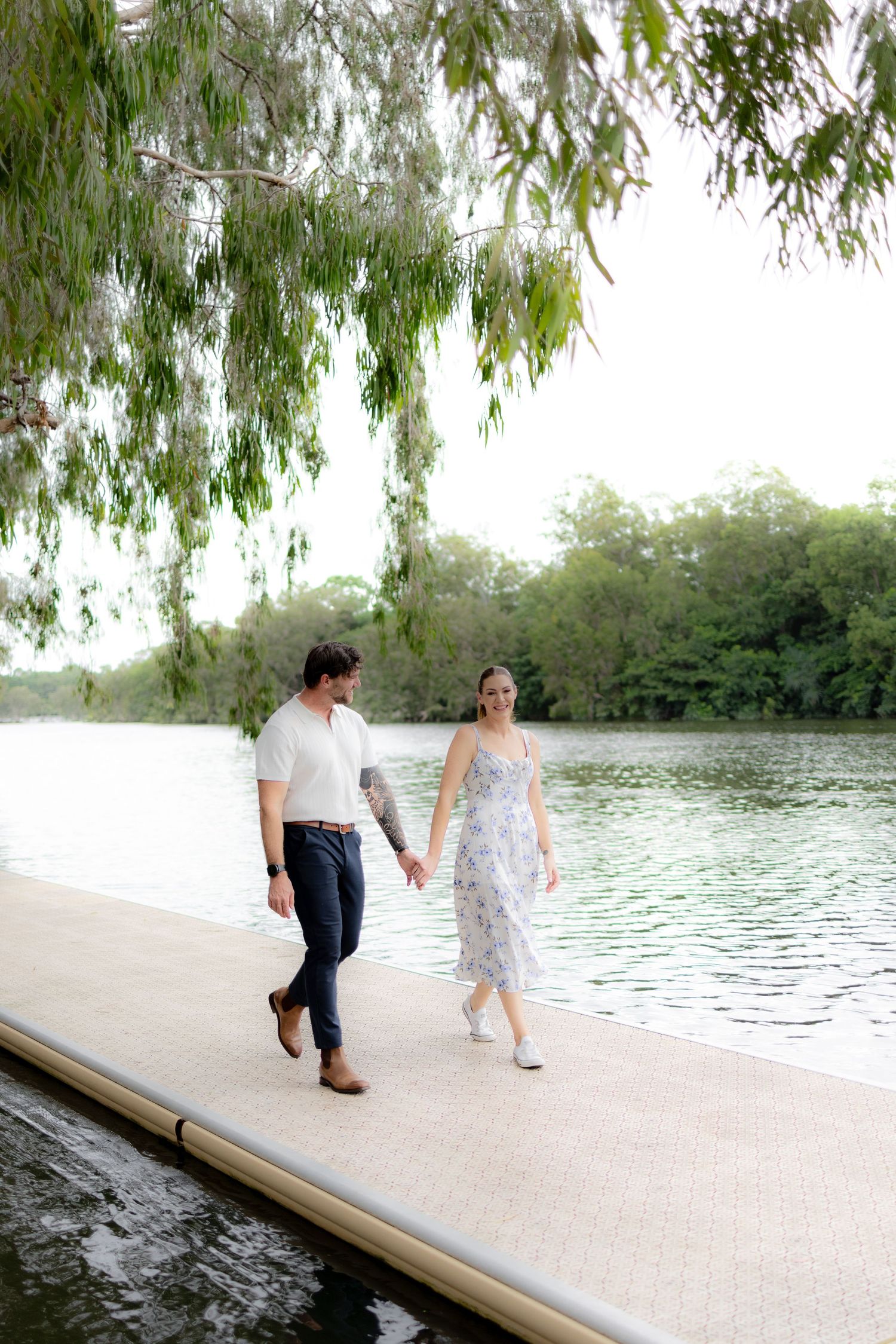 A couple walks along a tranquil lakeside path under overhanging willow trees on a sunny afternoon.