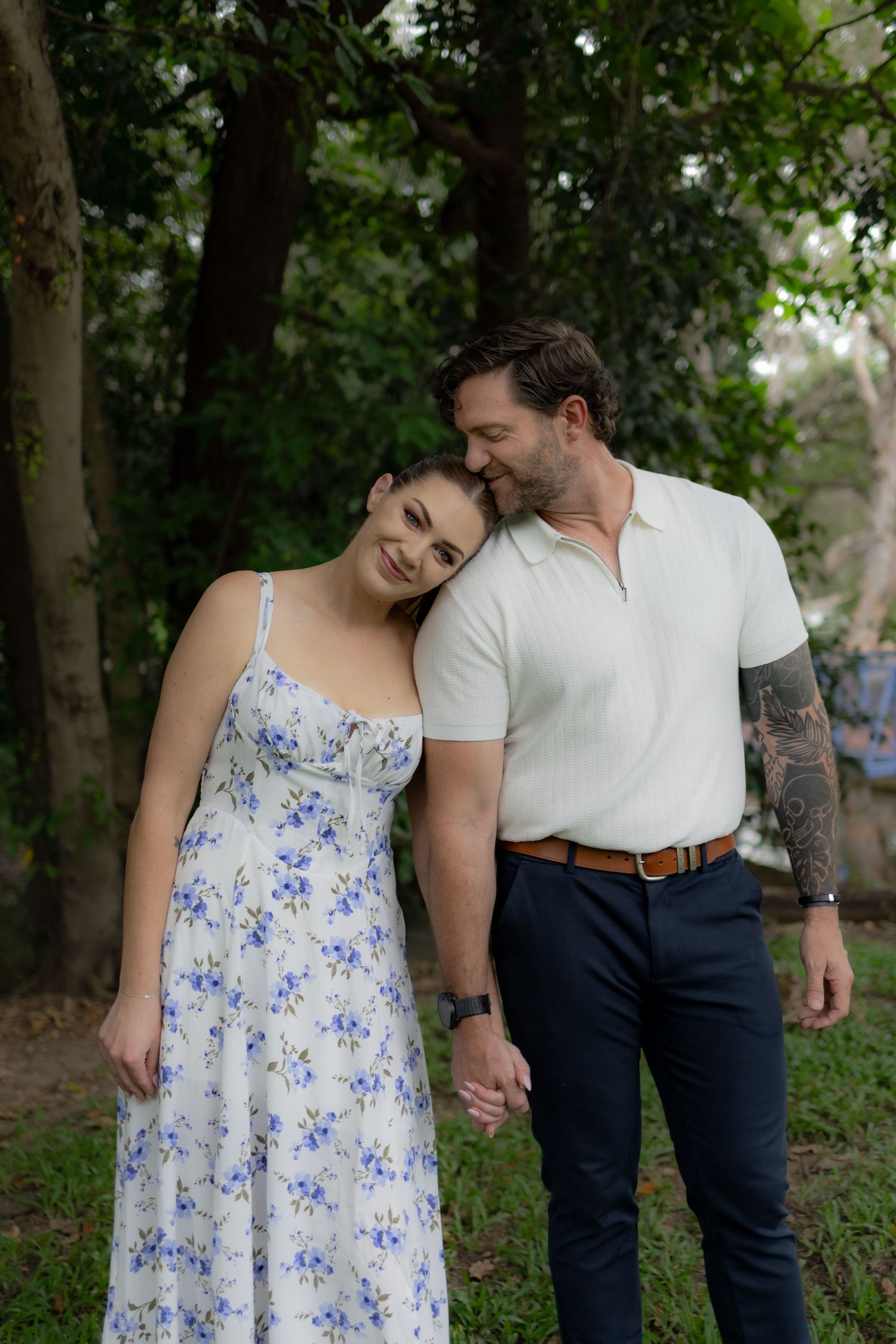 A couple shares a tender moment outdoors wearing a floral dress and casual attire against a wooded backdrop.
