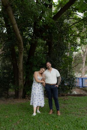 A tender moment between a couple in coordinated outfits standing together on green grass under tall trees.