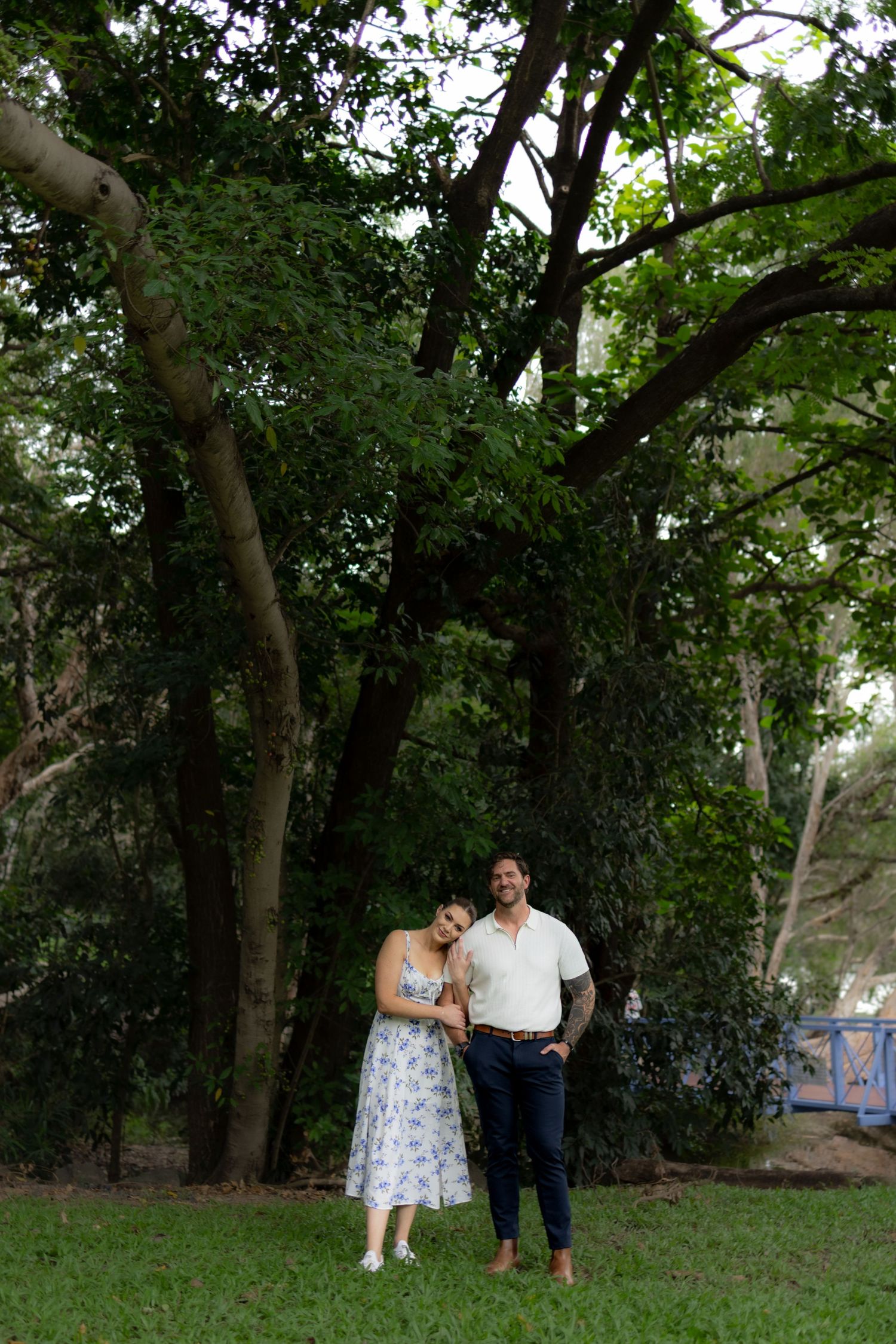 A couple shares a romantic moment under lush green trees in a wooded outdoor setting.