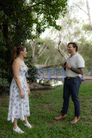 A couple shares special moments on grassy grounds with trees and a blue bridge in the background.