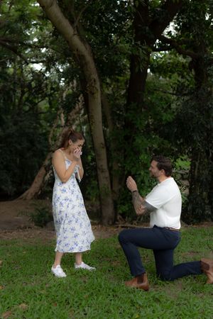 A couple shares a playful moment while sitting together on grass in a wooded area.