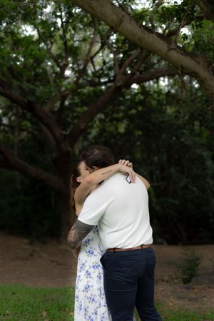 An intimate embrace between a couple in a floral dress and white shirt beneath towering trees.