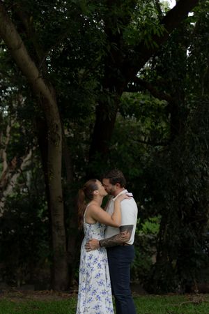 A romantic couple shares a tender embrace while standing in a wooded outdoor setting at dusk.