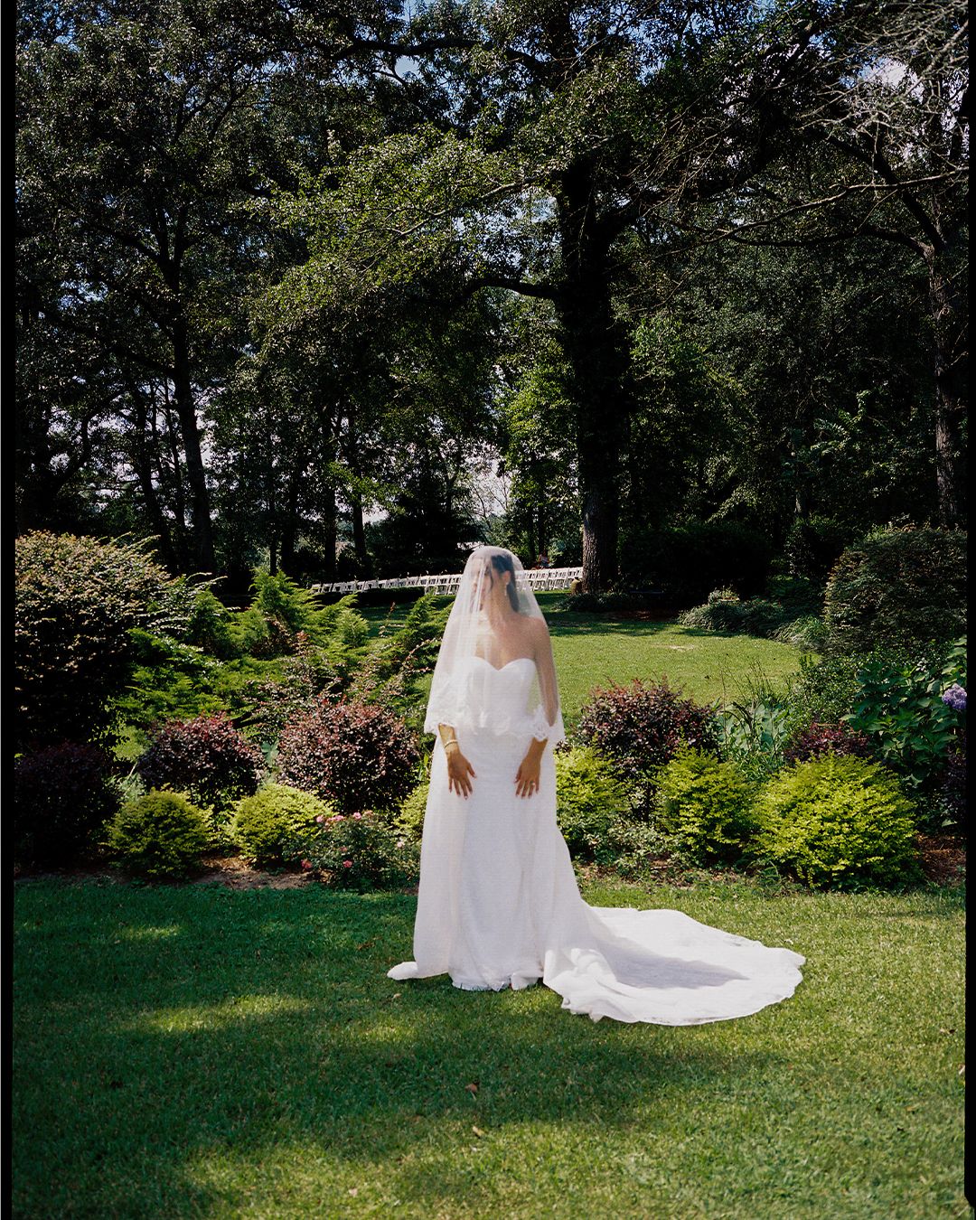 A bride in a flowing white wedding dress and long veil poses gracefully in a lush garden setting on a sunny day.