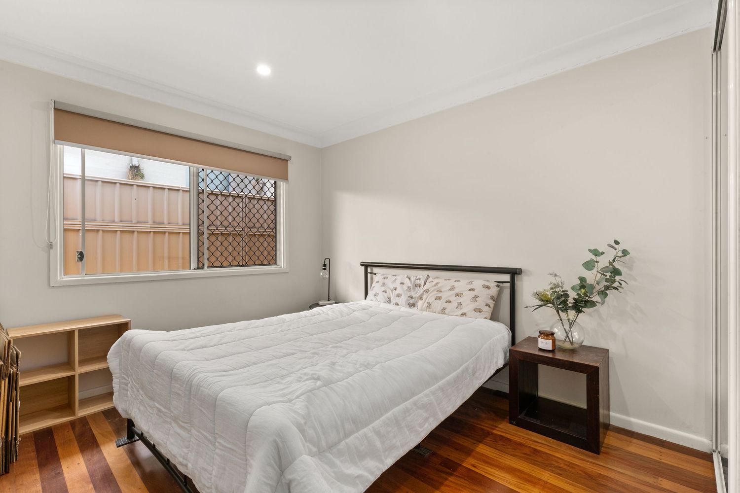 Simple bedroom with polished hardwood floors, white bedding, roller blind window, and timber bookshelf.