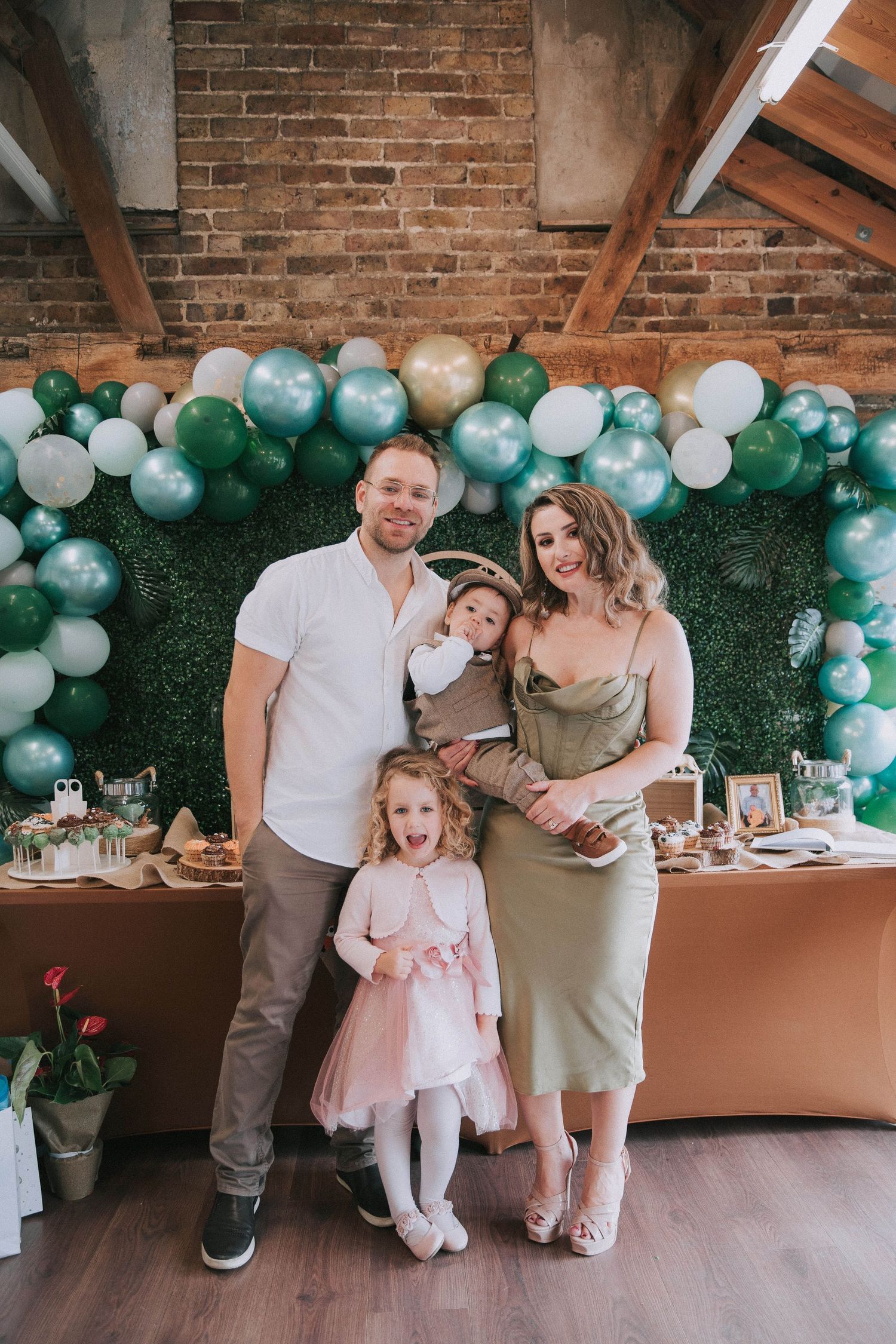 Family poses in front of balloon arch and dessert table at celebration with green and blue party decorations.