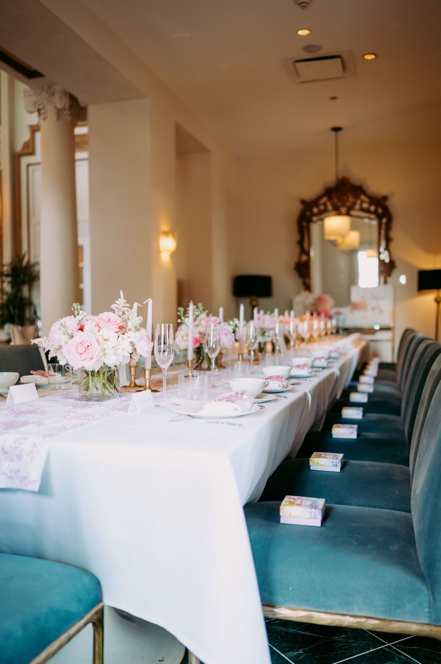 Elegant wedding reception table with pink floral centerpieces and teal velvet chairs along white linen tablecloth.