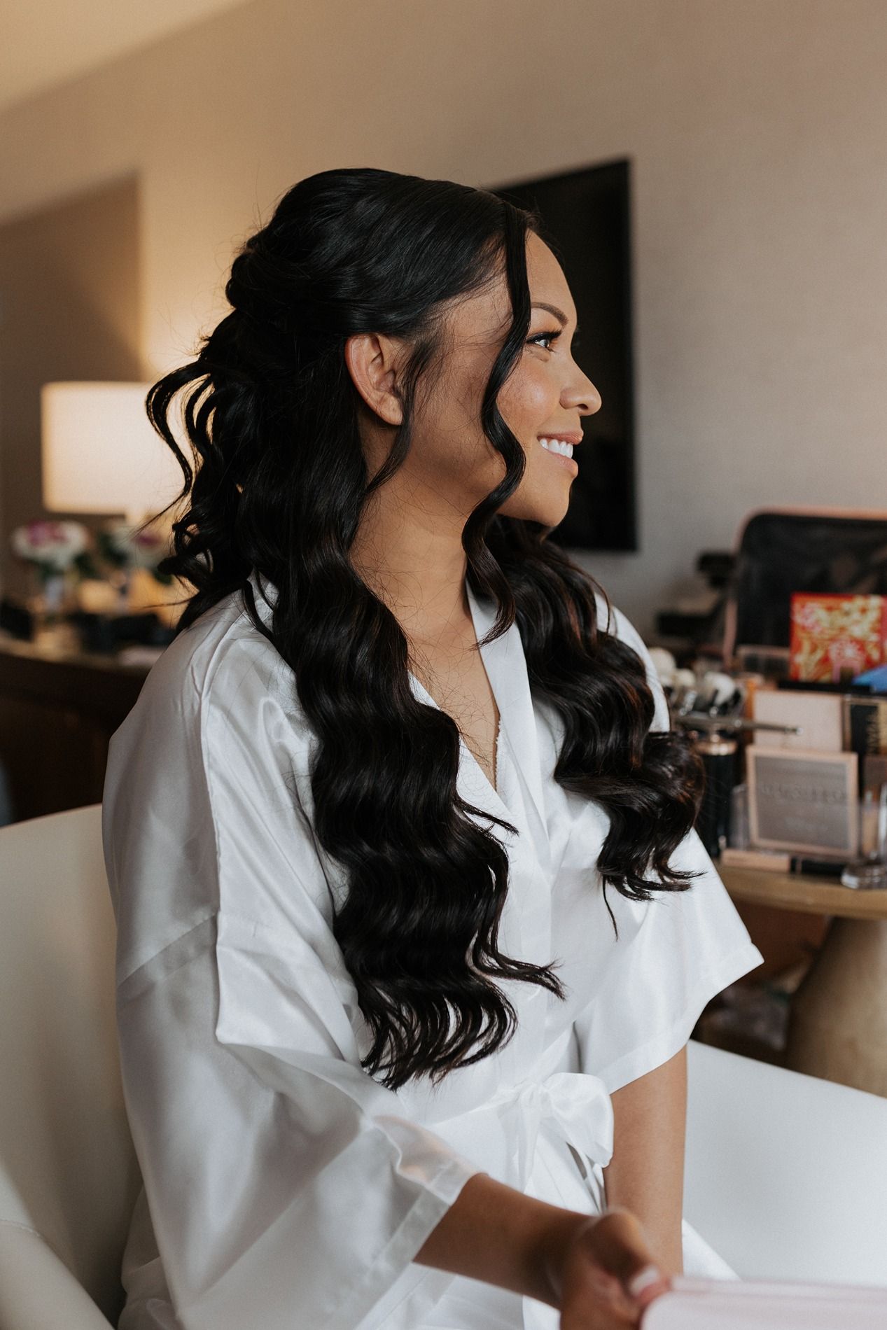 A bride with long dark wavy hair and a white robe while looking to the side in an indoor setting.
