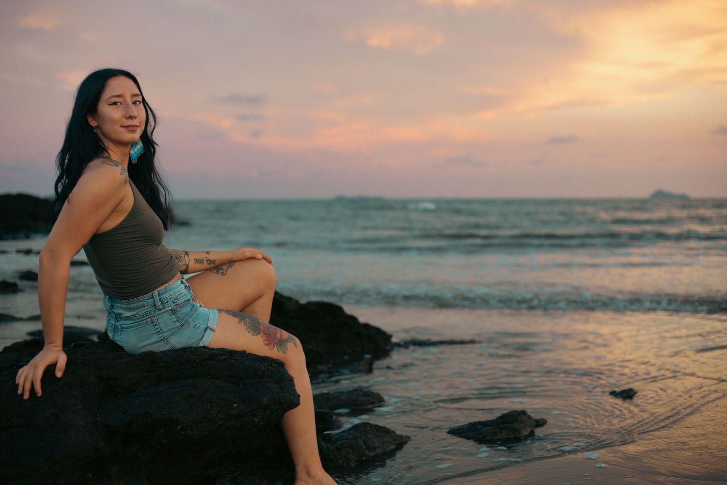 Woman sitting on rocks at beach during colorful sunset, wearing denim shorts and gray top, smiling peacefully.
