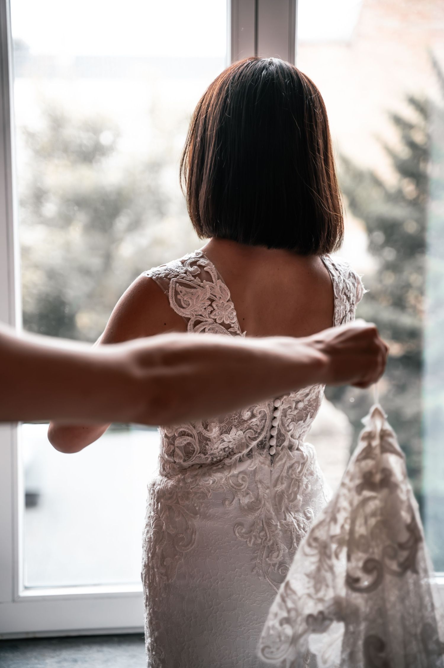Bride in intricate lace wedding dress being buttoned up near a bright window.