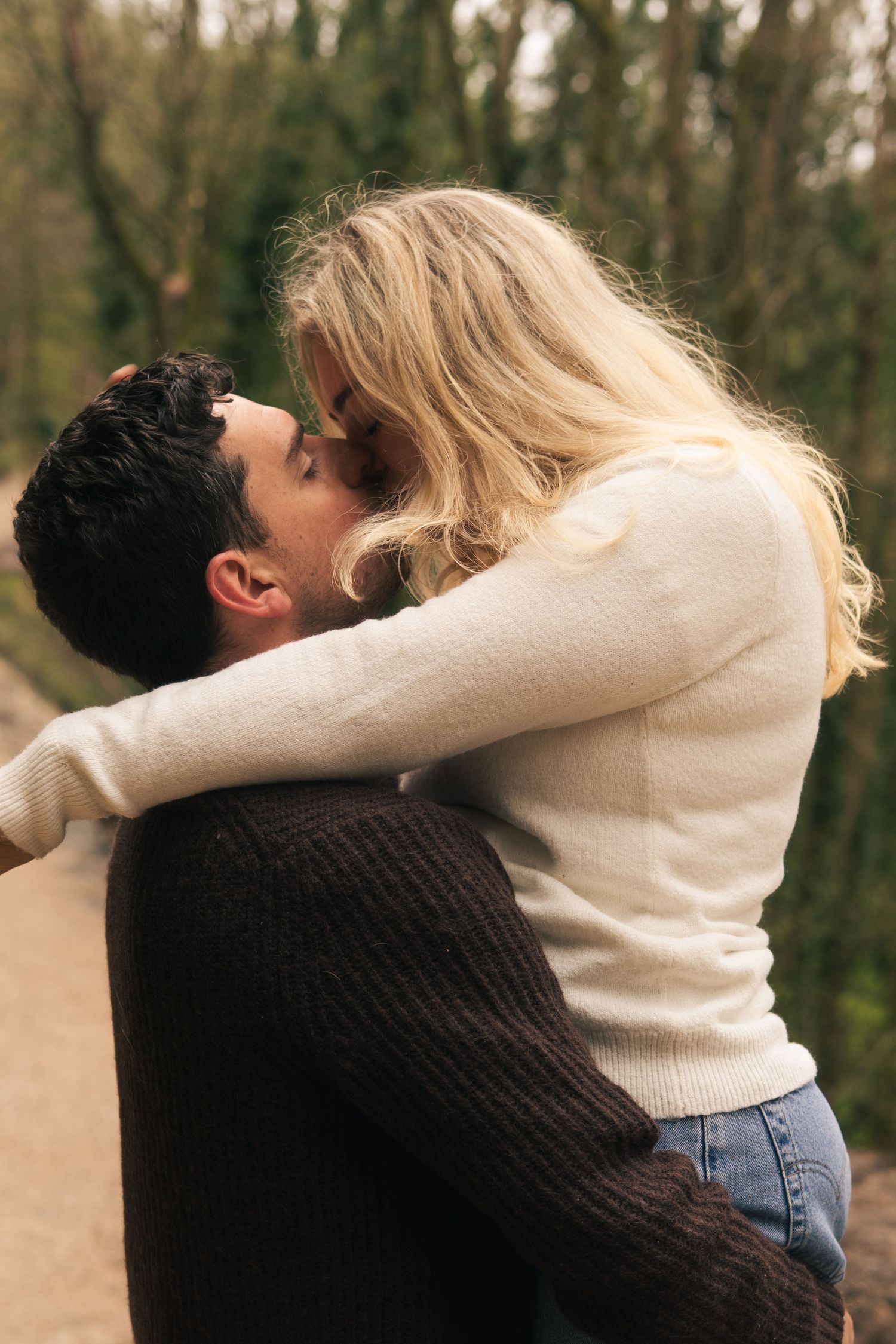 Couple sharing a tender kiss outdoors, woman with blonde hair embracing man in brown sweater among green trees.