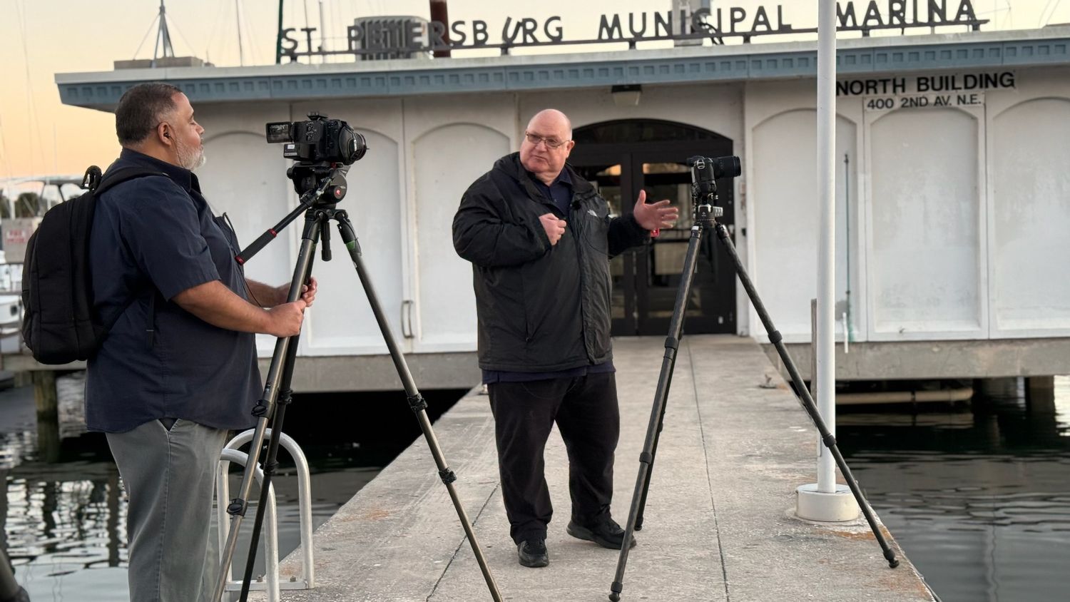 Two cameramen filming at St. Petersburg Municipal Marina on a wooden dock at dusk.