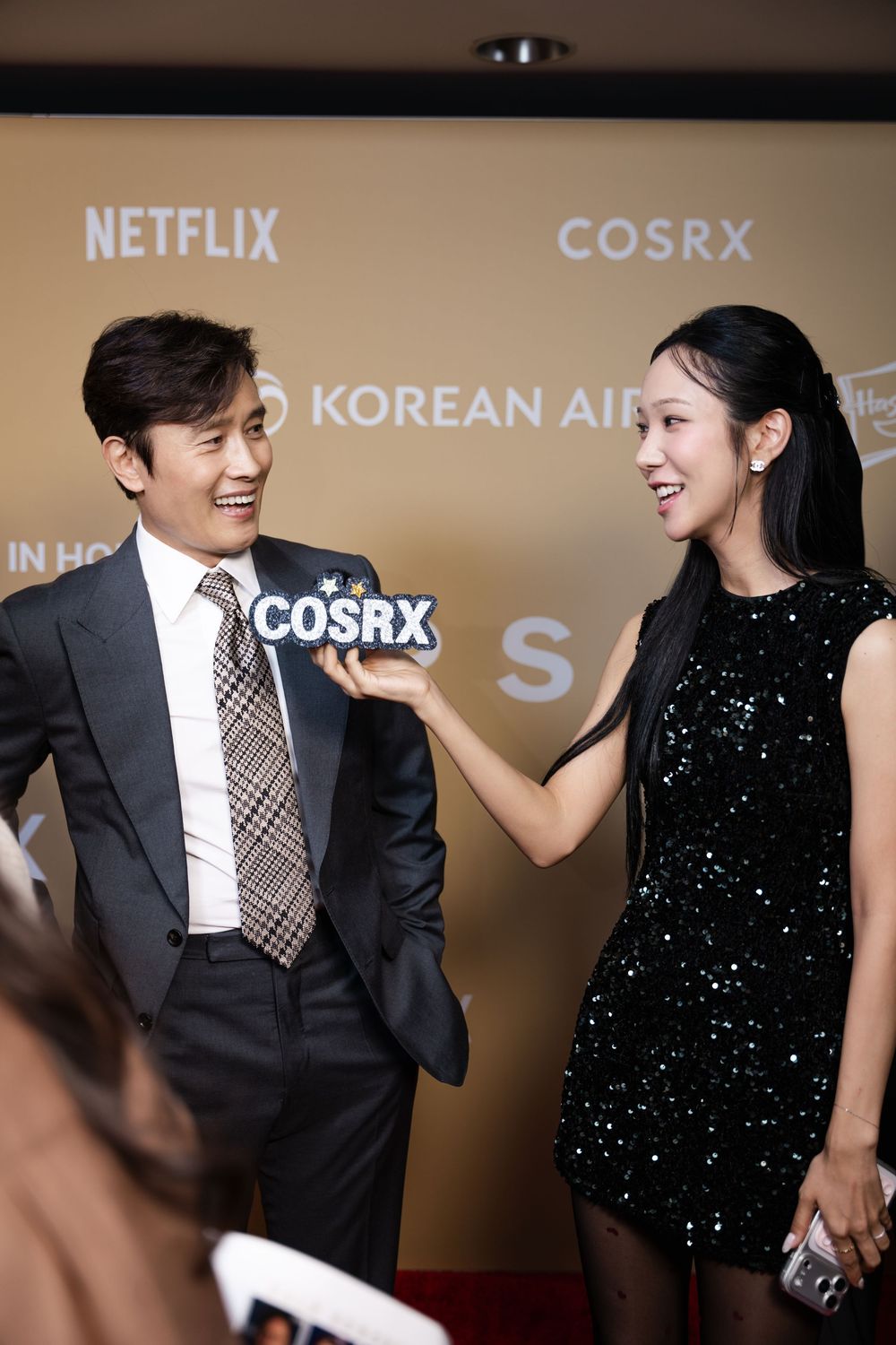 Two elegantly dressed attendees pose at a Netflix and COSRX Korean Air red carpet event, holding a COSRX branded sign.