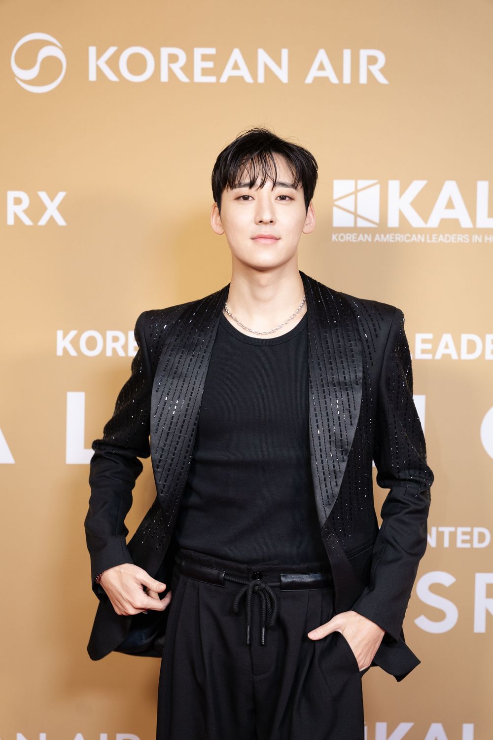Man in black sequined blazer posing at Korean Air and KALRX event on gold backdrop.