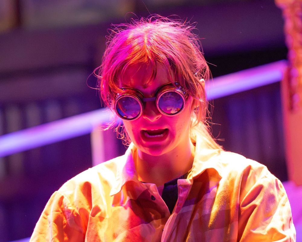 A performer in pink lighting dances energetically on stage wearing yellow clothing and round glasses.