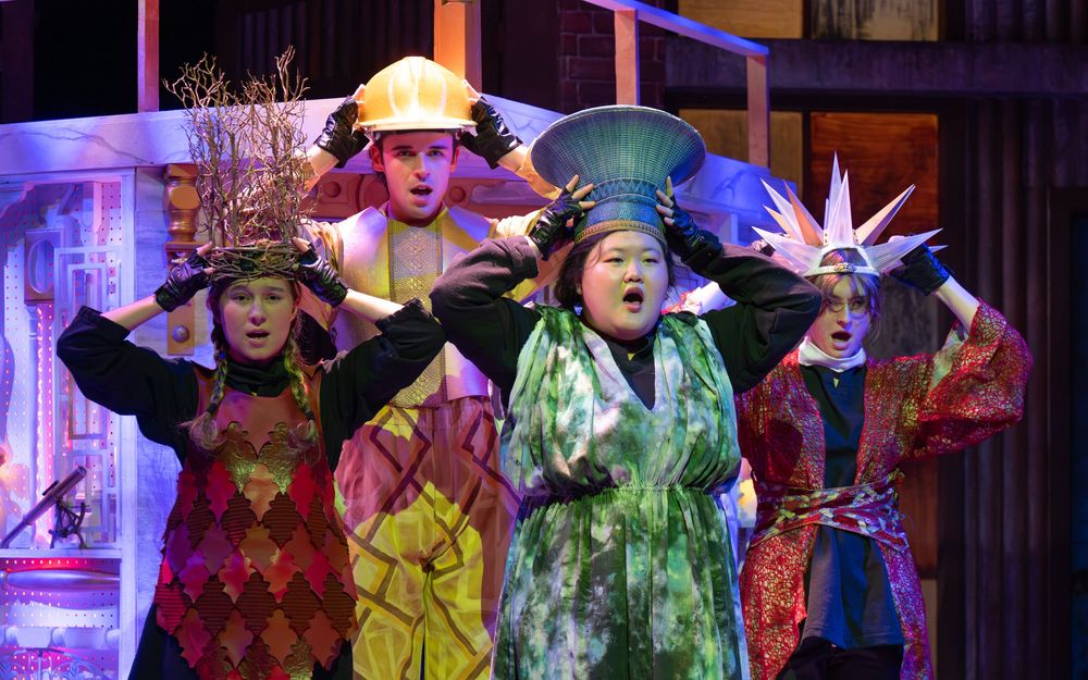 Performers in colorful costumes dance and pose during a theatrical stage production with vibrant lighting effects.