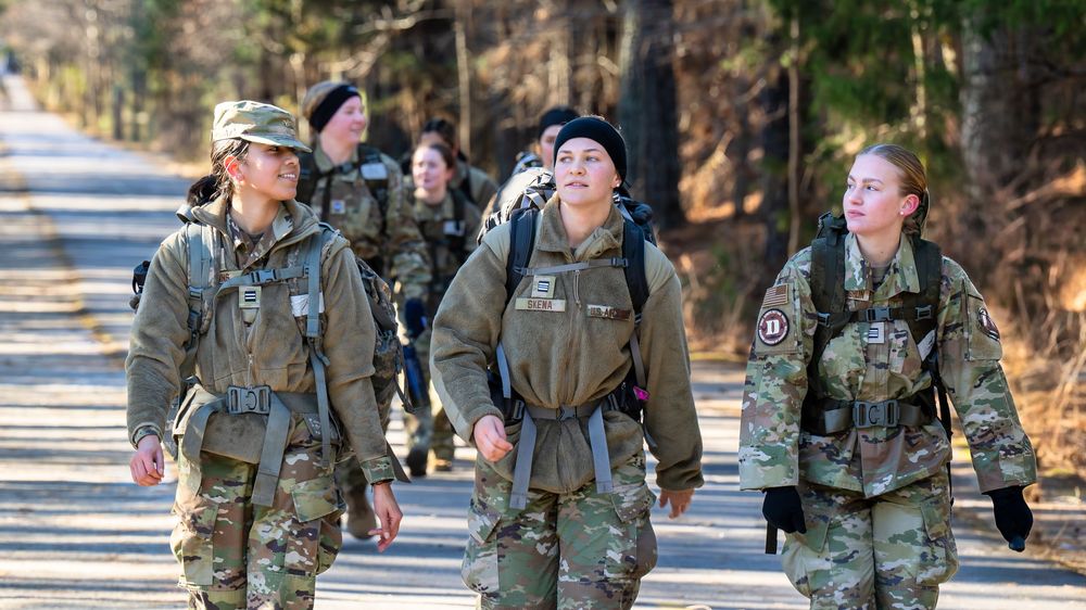 Military personnel in camouflage uniforms walk in formation along a paved road during training exercises.