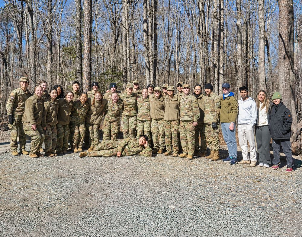 Large group of military service members in camouflage uniforms standing together outdoors in a wooded area.