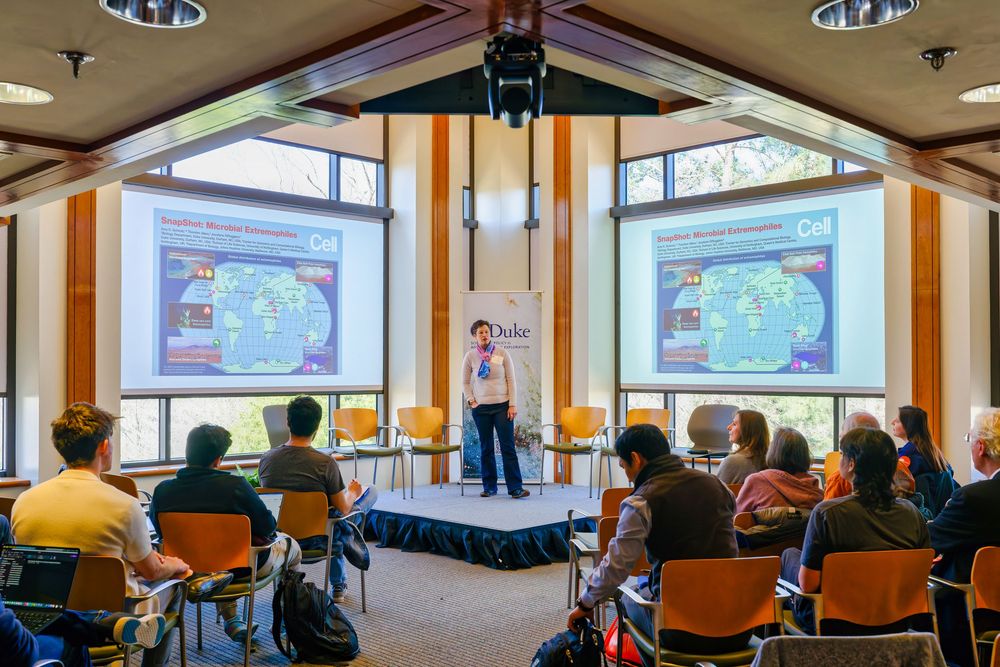 Students attend a lecture in a modern classroom with multiple projector screens displaying educational content.