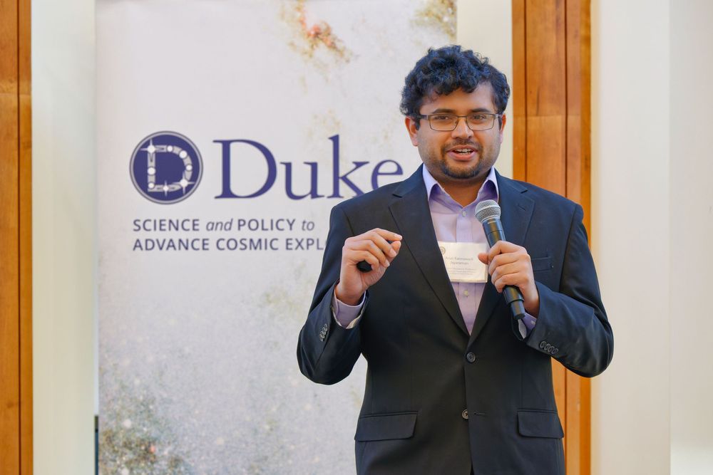 A person in a dark suit gestures enthusiastically while speaking at a Duke University podium.