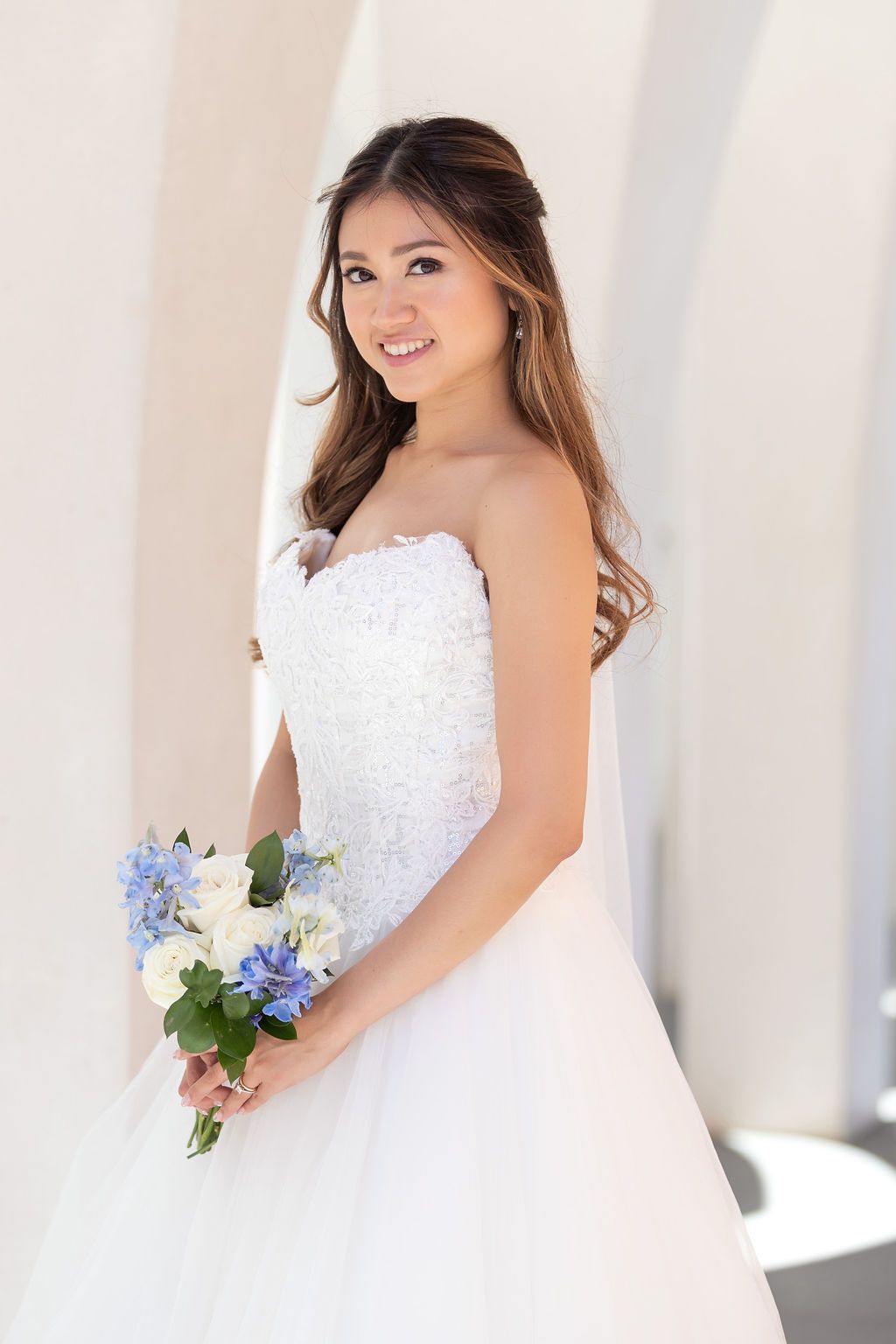 Smiling Asian bride in strapless ballgown holds blue and white floral bouquet outdoors.