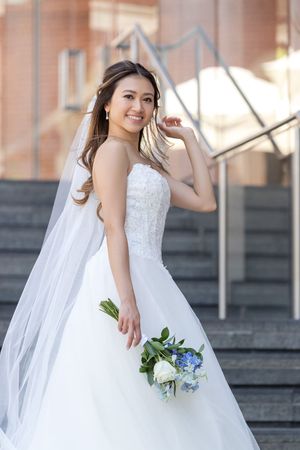 Radiant Asian bride in strapless ballgown poses on steps with blue and white floral bouquet.