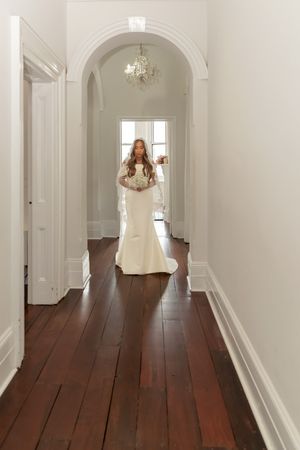 Bride in a fitted white gown holding a bouquet walking through an elegant hallway with arched doorway.