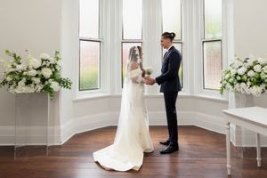 Bride and groom holding hands during an intimate wedding ceremony by tall windows with white floral arrangements.