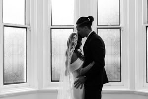 Black and white romantic photo of bride and groom embracing tenderly in front of large windows.