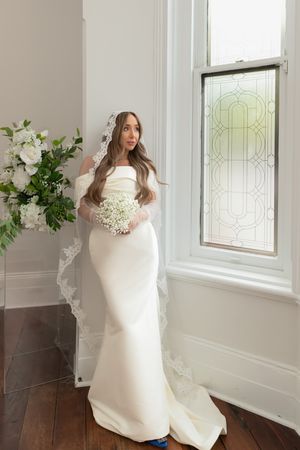 Beautiful bride in a fitted white gown holding a white bouquet standing by a stained glass window.