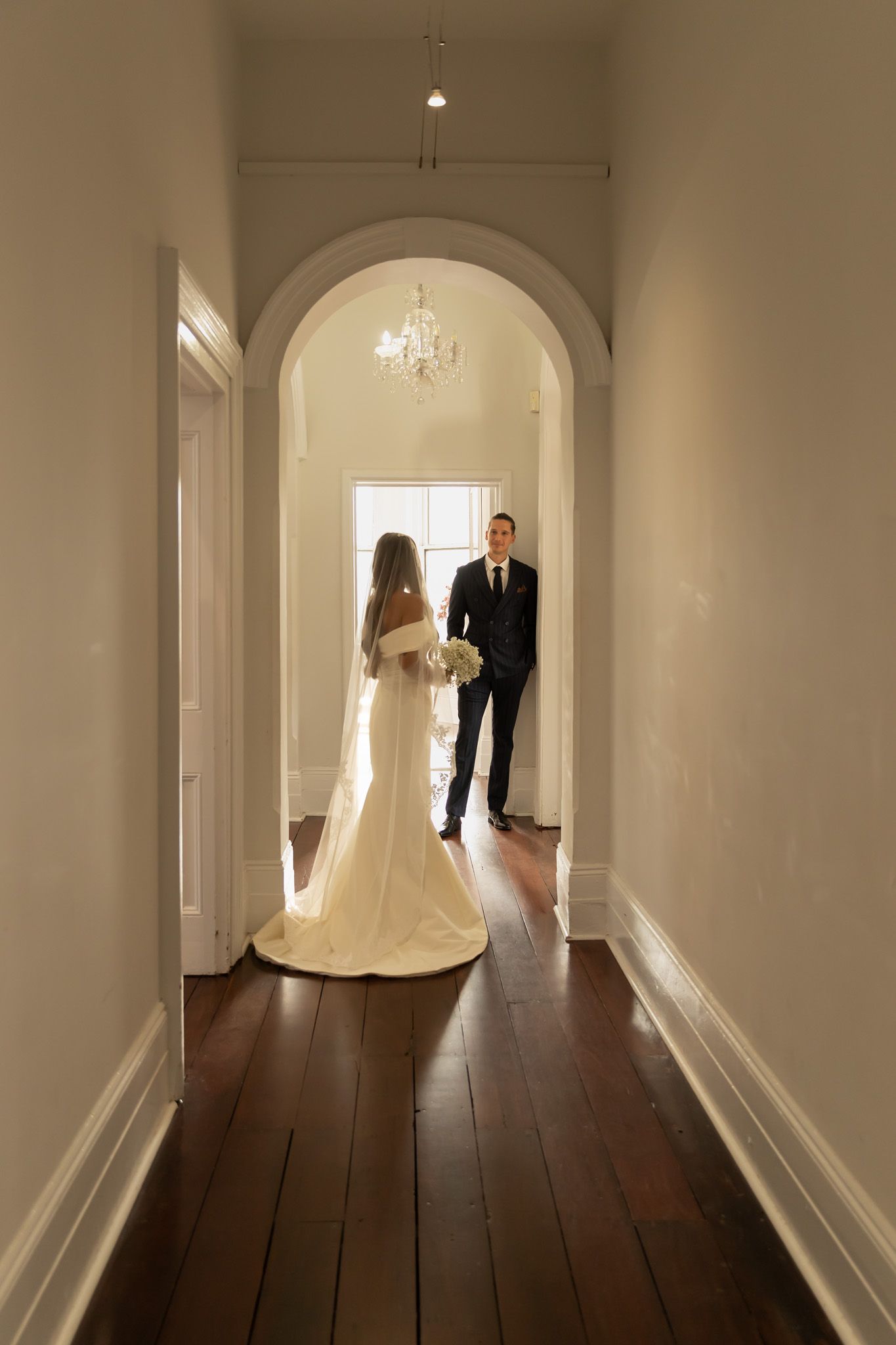 Bride in elegant white gown and veil walks toward groom in a chandelier-lit hallway during wedding first look.