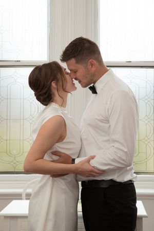 Couple in wedding attire share intimate moment near frosted glass window indoors.