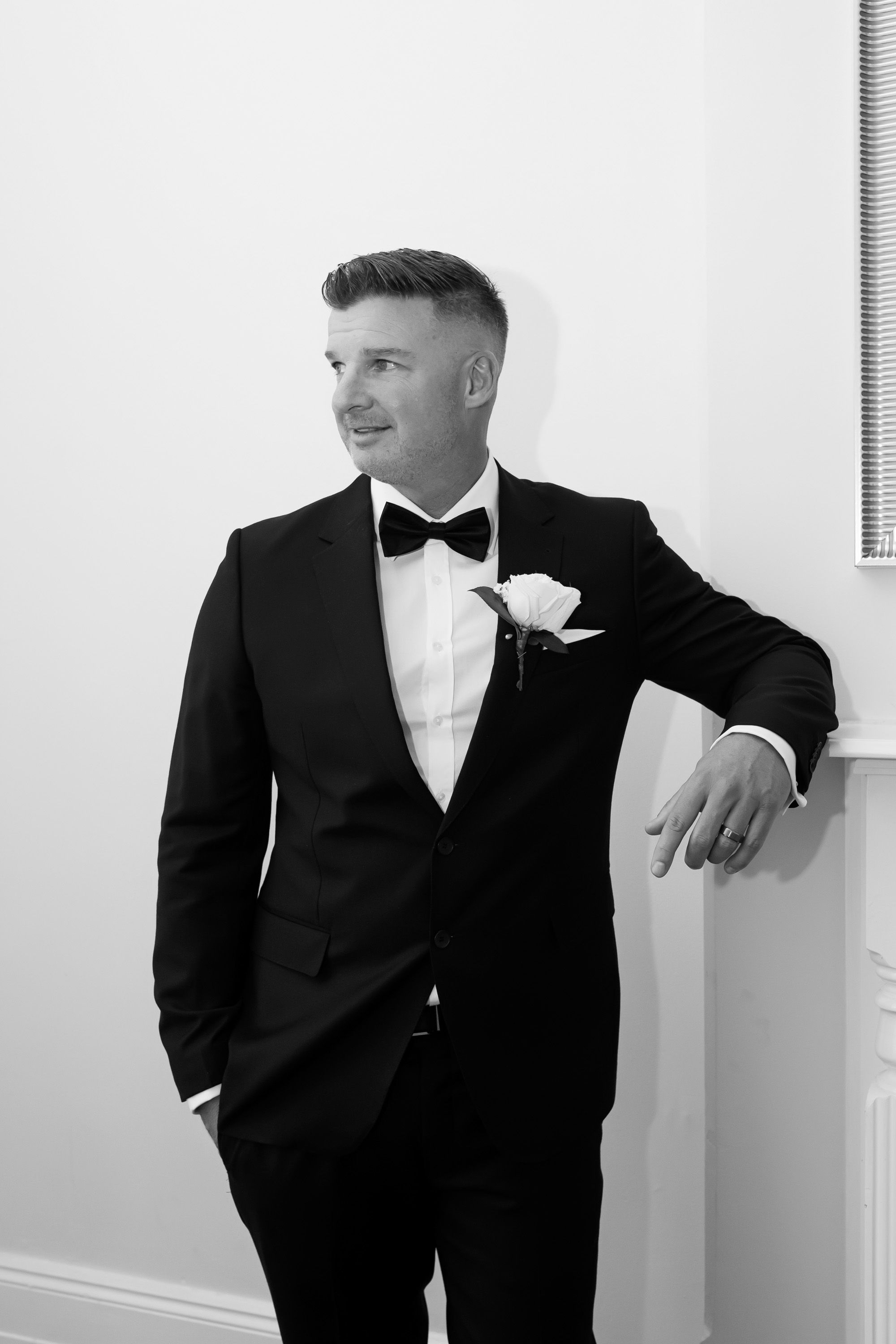 Elegant groom in black tuxedo with bow tie and rose boutonniere poses against a white wall in black and white.