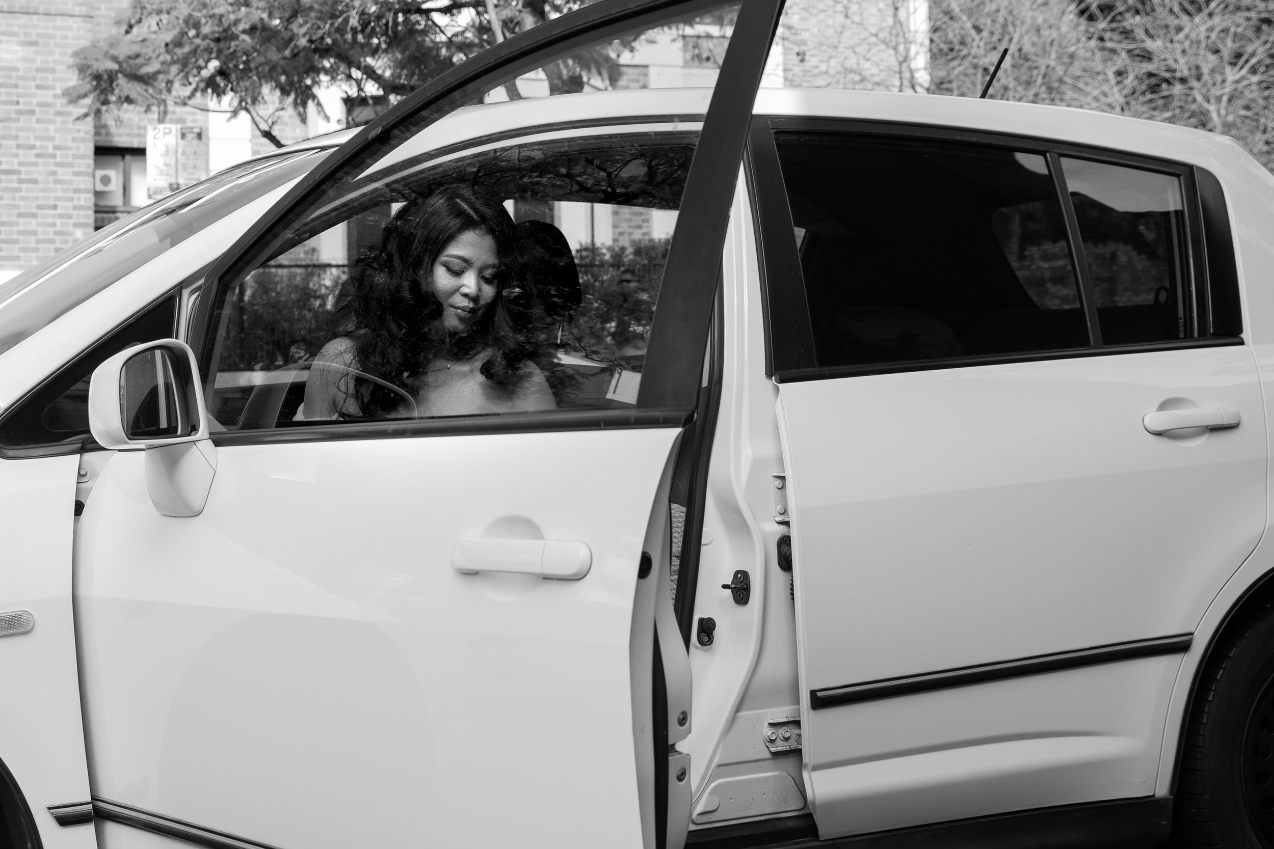Bride in white wedding dress smiles while seated in a white car in a classic black and white photo.