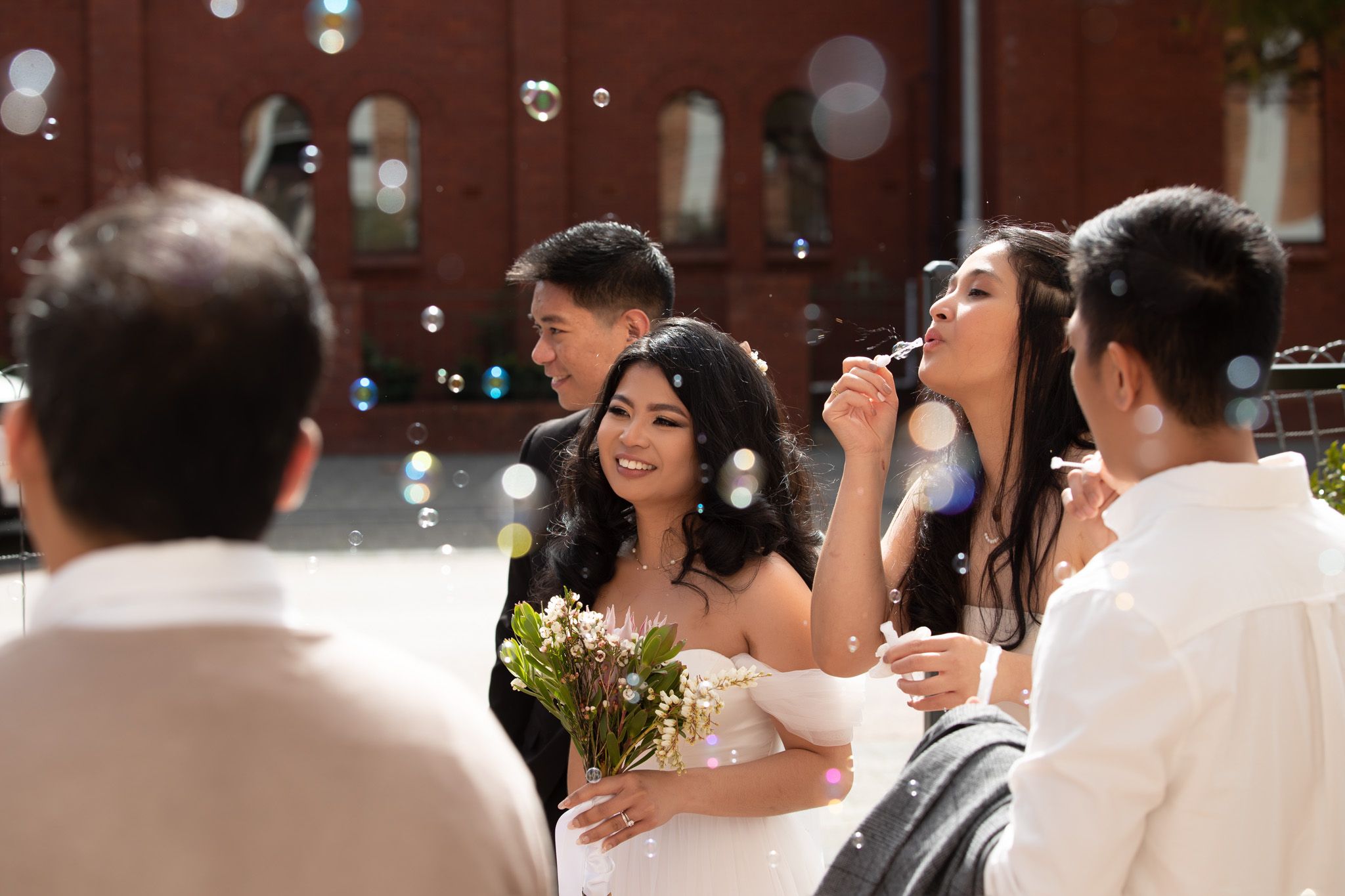 Smiling bride holding bouquet blows bubbles with guests outside brick building wedding celebration.