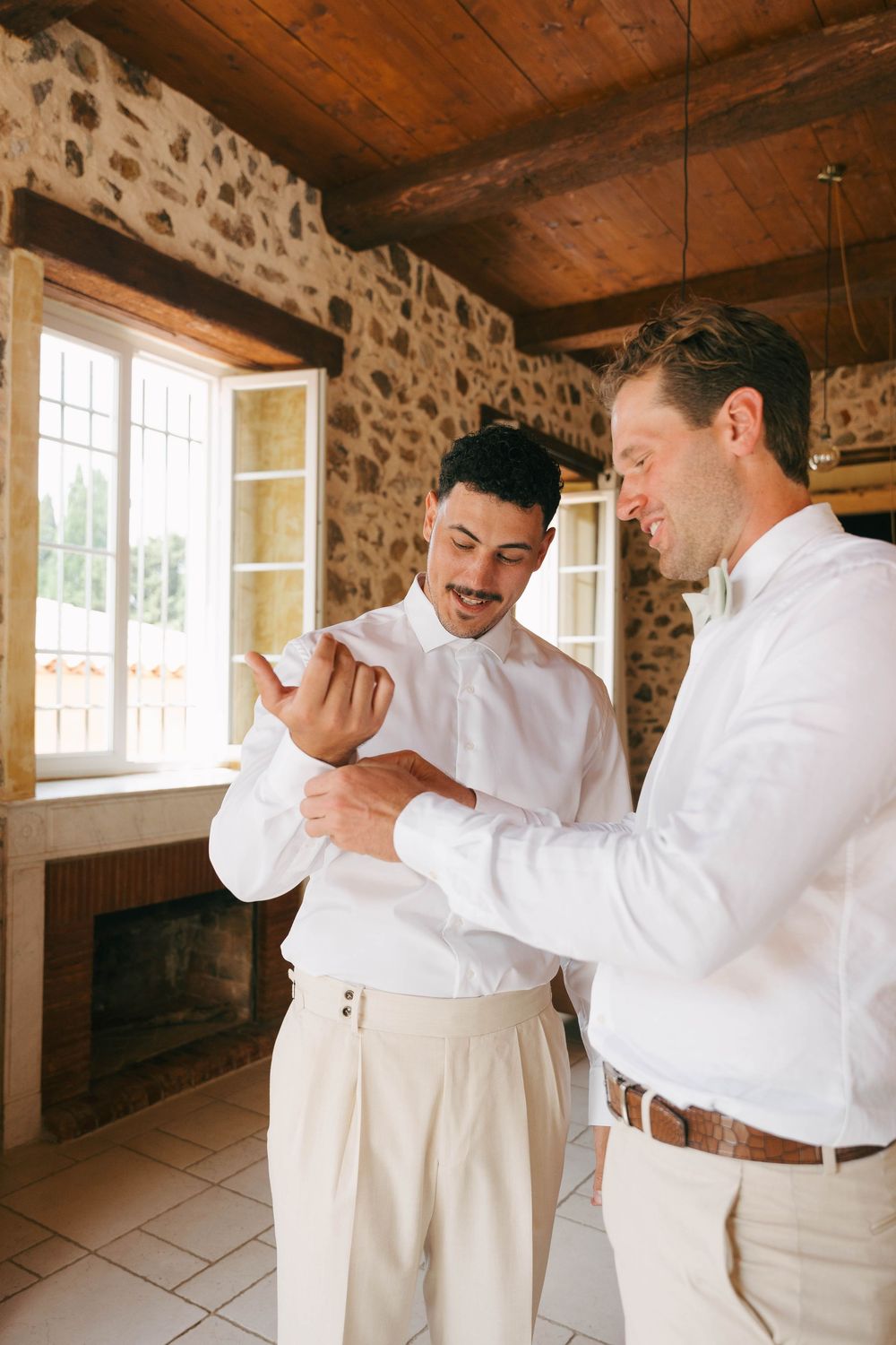 Two people in white shirts and beige pants share a candid laughing moment in a rustic stone-walled room.