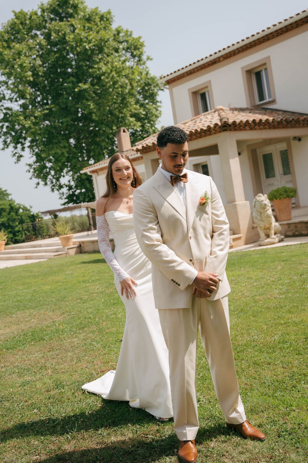 A couple in matching white outfits poses playfully on a grassy lawn in front of a Mediterranean-style building.