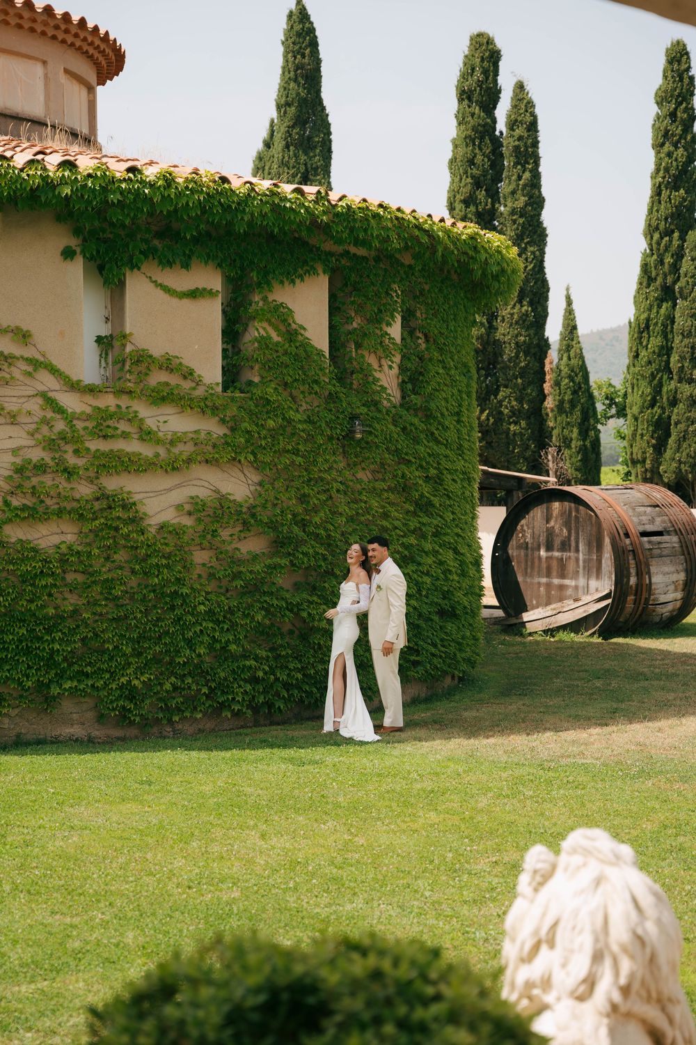 A couple stands together on a lush green lawn beside an ivy-covered wall at a Mediterranean-style venue.