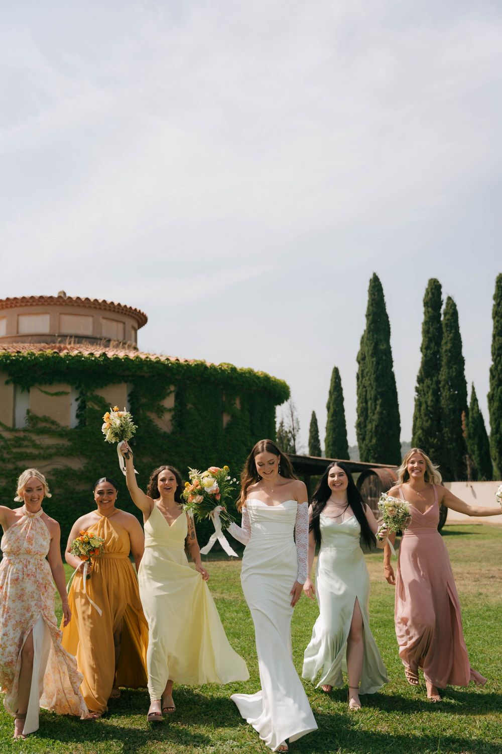 A line of bridesmaids in pastel and yellow dresses walk together on a lawn with cypress trees in Tuscany.